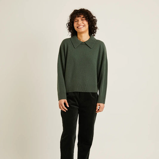 A woman modeling a dark green long-sleeve knit sweater with a collar, paired with black trousers and black flats, against a plain light gray studio background.