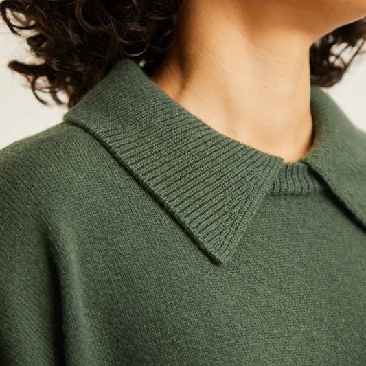 Close-up of a person wearing a dark olive green knit sweater with a wide ribbed collar, showing the neck, hair, and upper chest against a plain light background.