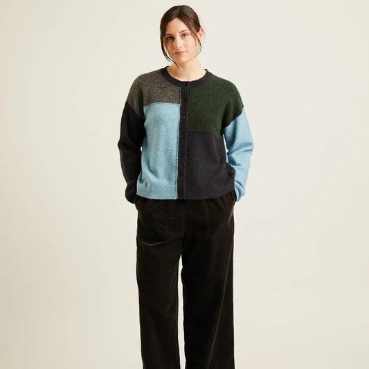 A woman stands against a plain light beige studio background wearing a color-block knit sweater with dark green, light blue, navy, and gray panels, black wide-leg corduroy pants, and black flats.