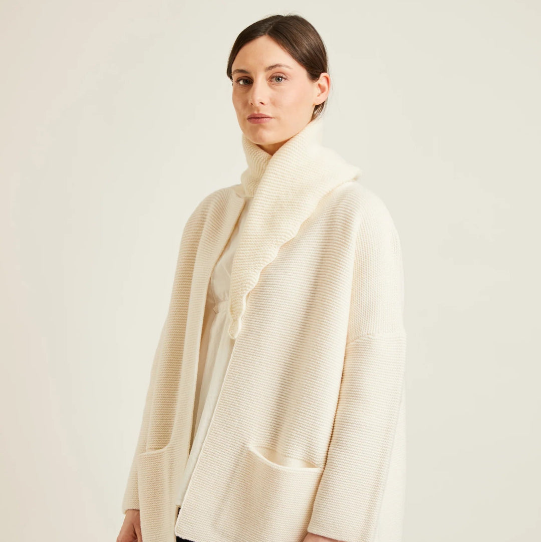 A woman modeling a cream-colored textured knit cardigan and a matching scalloped scarf, paired with black trousers, against a plain light beige studio background.
