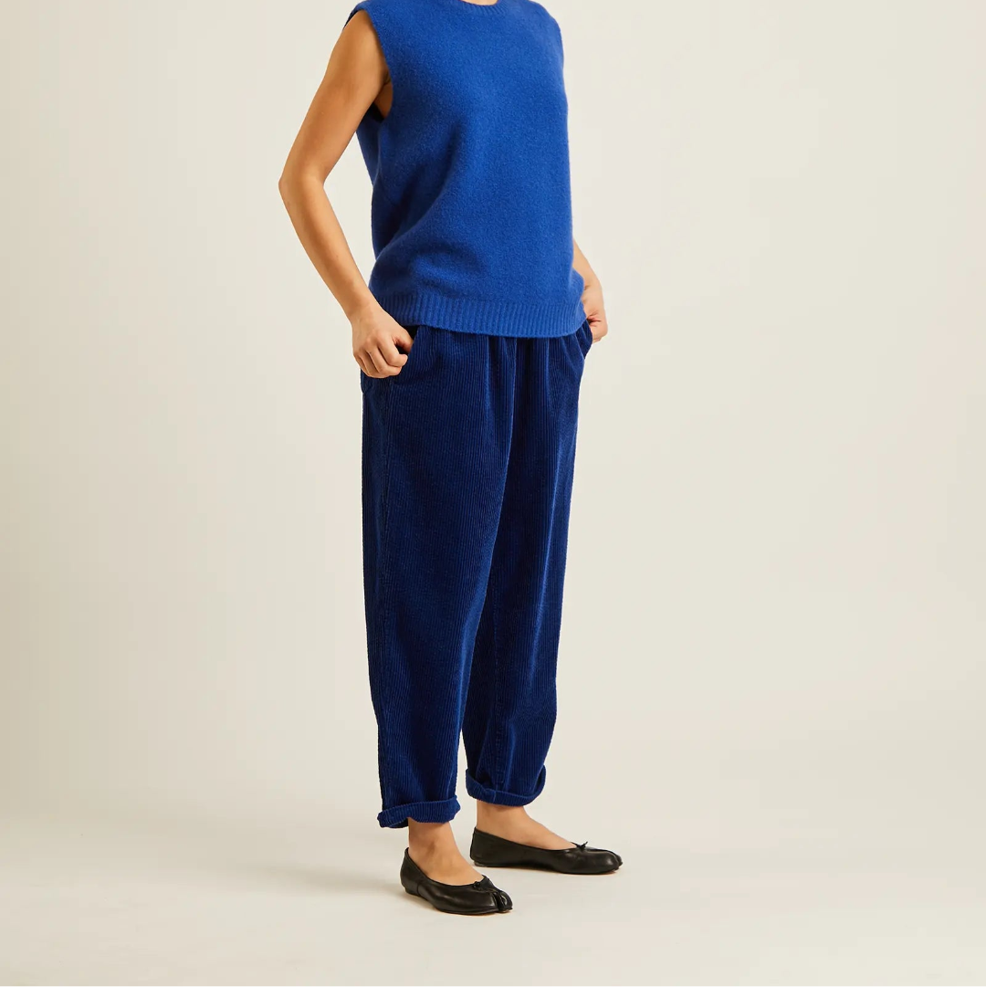 A model wearing a vibrant blue sleeveless knit sweater vest and matching blue velvet jogger pants, paired with black flats, standing against a plain light beige studio background.