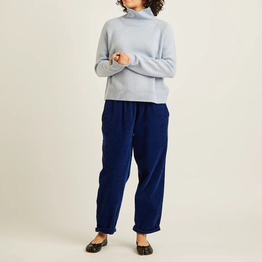 A model wearing a light blue turtleneck sweater and navy blue corduroy pants, standing against a plain off-white studio background.