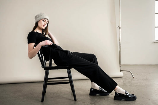 A model wearing a black t-shirt, black staight-leg trousers, white socks, and black loafers sits on a black wooden chair in a minimalist studio setting.