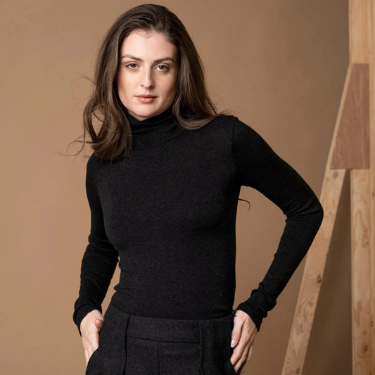 A woman modeling a black long-sleeve turtle neck knit top and a black pleated skirt against a warm brown studio backdrop with a wooden structure on the right.