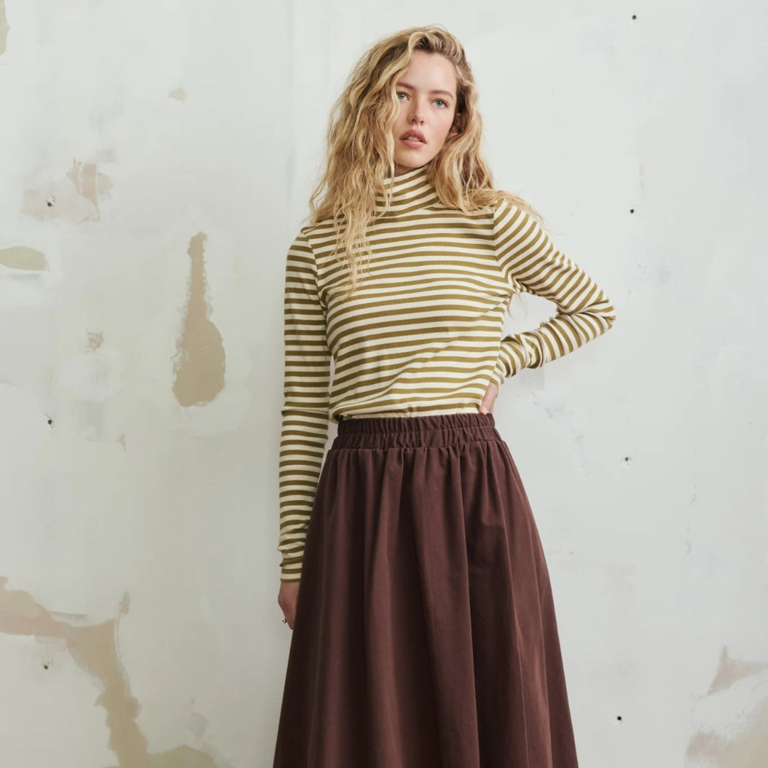 A woman stands against a textured off-white wall wearing a olive green and white striped mock-neck long-sleeve top and a dark brown pleated maxi skirt, paired with dark brown slip-on shoes.