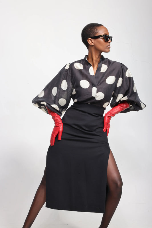 A model wearing a black midi skirt with a thigh-high slit, a sheer black blouse with large white polka dots and dramatic elbow-length sleeves, bright red gloves, and black sunglasses, posed against a light gray studio background.