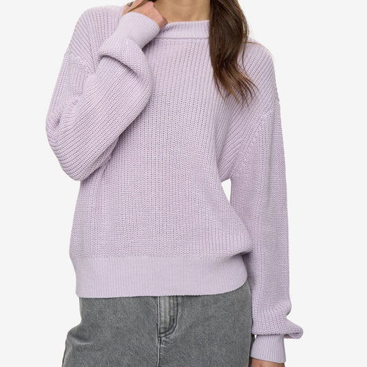 A woman wearing a lilac ribbed knit sweater and gray jeans, posing against a solid white studio background.