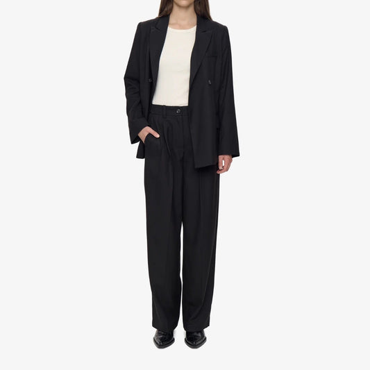 A model stands against a white studio background wearing a black double-breasted blazer and matching wide-leg trousers over a plain, white top.