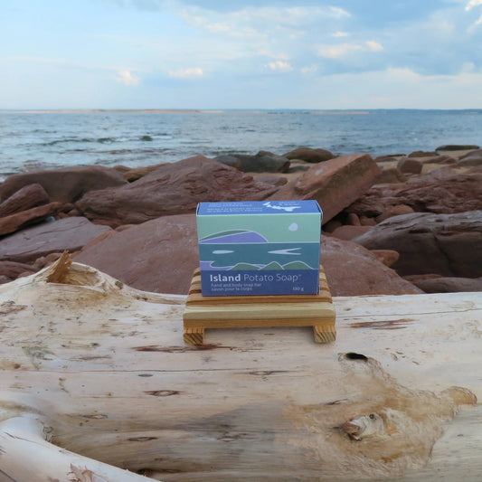 A small box of Island Potato Soap sits on a wooden stand on a driftwood log, with a rocky shoreline and ocean in the background.