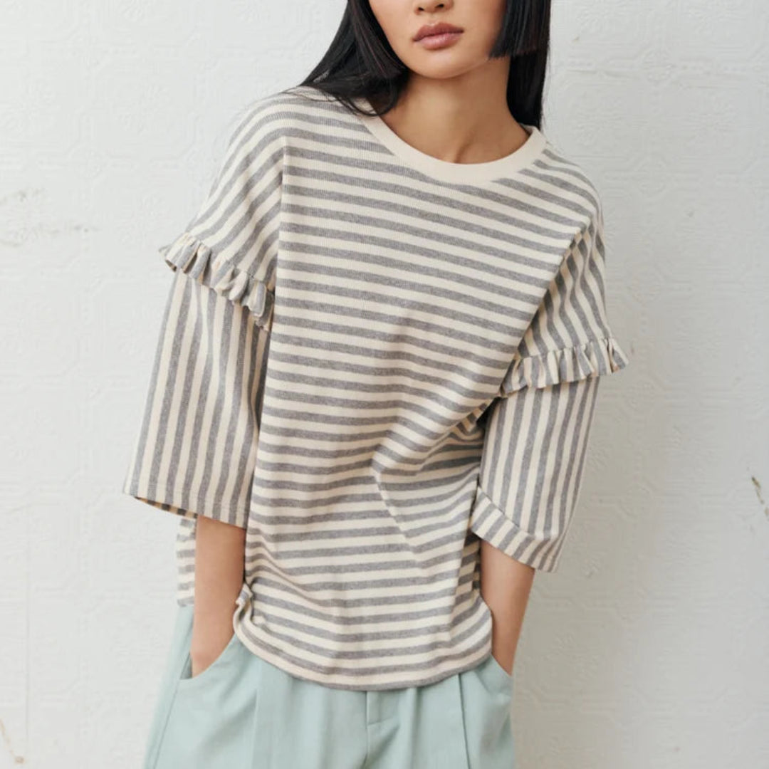 A woman with a straight black hair wearing a cream and gray striped ruffle-sleeve top and light blue trousers, standing against a white textured wall.