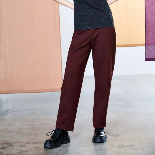 A woman modeling a short-sleeve black knit top and burgundy wide-leg trousers with black shoes, standing against a modern studio backdrop with textured panels.