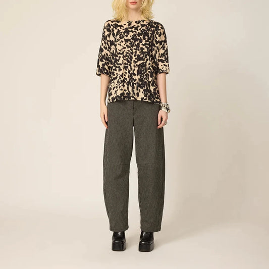 A blonde model stands against a plain light beige studio background wearing a short-sleeved leopard-print top with black and beige tones, dark gray and black striped barrel-leg trousers, and black platform shoes.