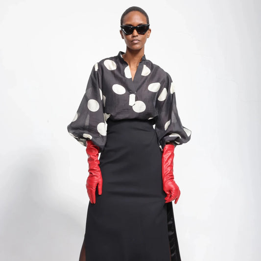 A model wearing a black sheer long-sleeve blouse with large white polka dots, a black midi skirt, and bright red gloves, posed against a plain white studio background.