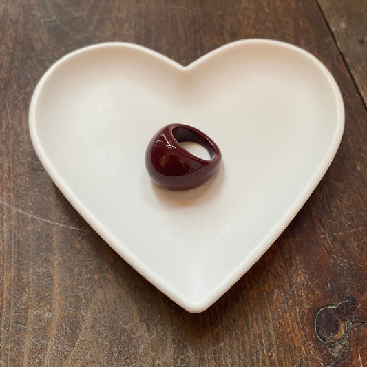 A glossy dark red ring displayed in the center of a white heart-shaped ceramic dish on a rustic dark wood tabletop.