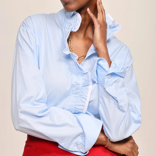 A model with short dark hair wearing a light blue long-sleeve blouse with ruffled details and a red skirt, posed against a solid beige background.