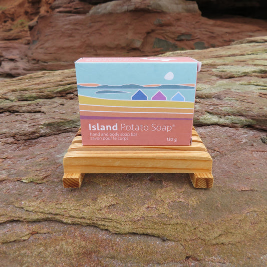 A boxed bar of soap labeled 'Island Potato Soap' sits on a small wooden stand outdoors against a rocky, reddish-brown natural background.