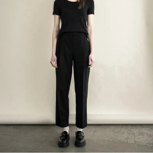 A model stands against a plain beige wall wearing a black short-sleeve top, black straight-leg trousers, and black shoes.