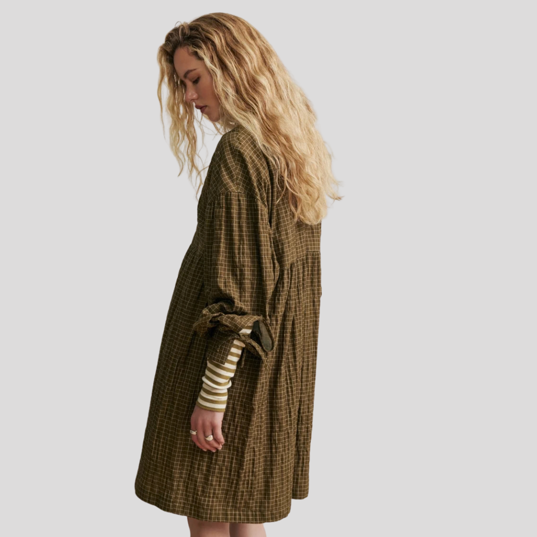 A woman with long, wavy blonde hair is shown from a side-back angle wearing a loose, oversized brown and olive green plaid dress over a cream and brown striped long-sleeve shirt, set against a solid light gray background.