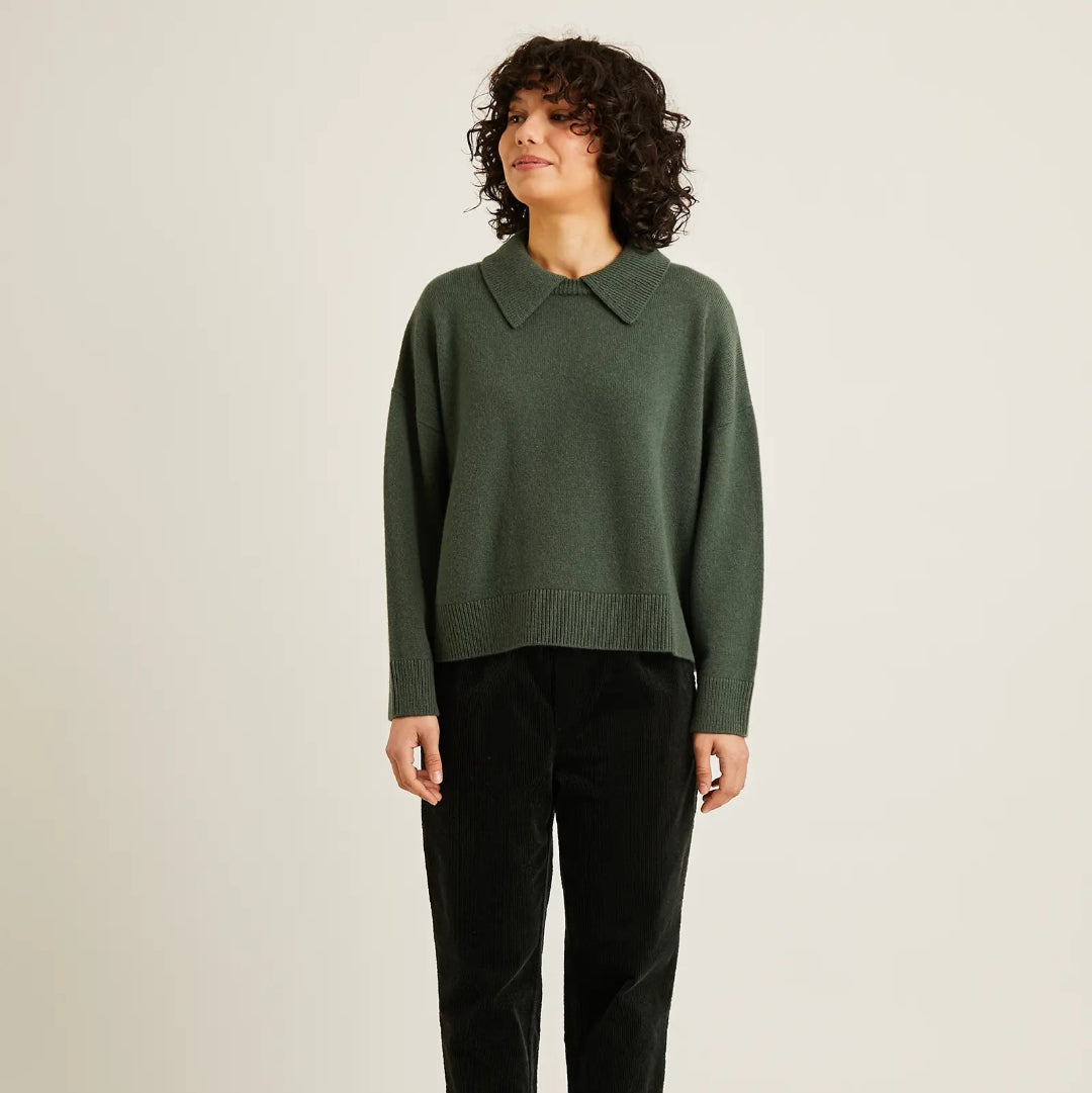 A woman stands against a plain light beige studio background wearing a dark green knit sweater with a pointed collar, black trousers, and black flats.