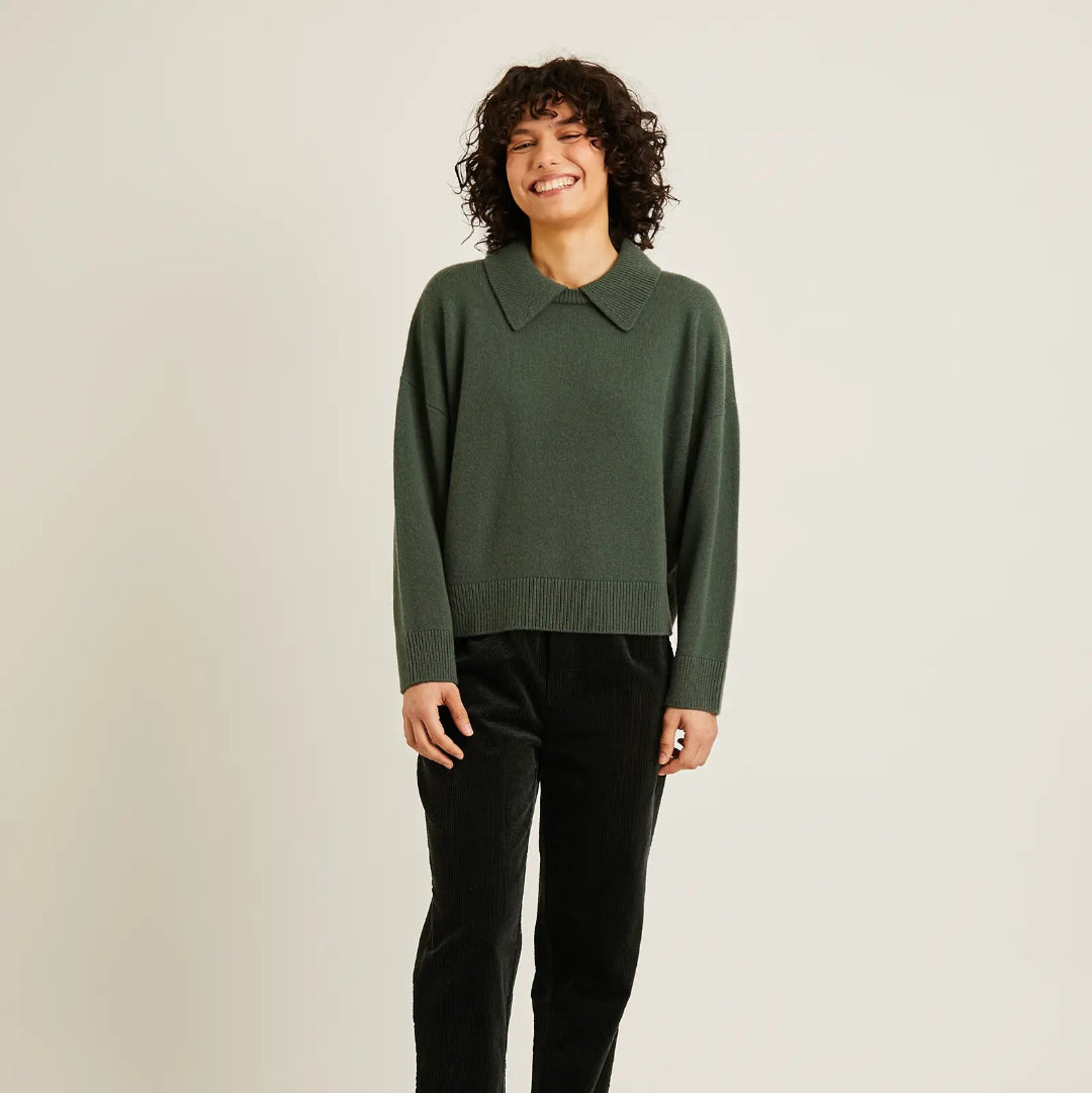 A woman modeling a dark green long-sleeve knit sweater with a collar, paired with black trousers and black flats, against a plain light gray studio background.
