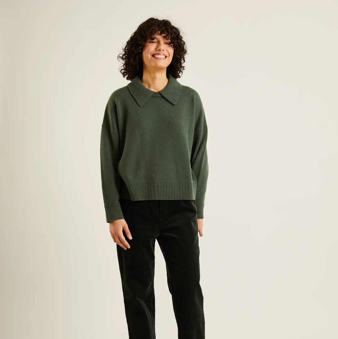 A woman stands against a plain light beige studio background wearing a dark green long-sleeve knit sweater with a pointed collar, black trousers, and black flats.