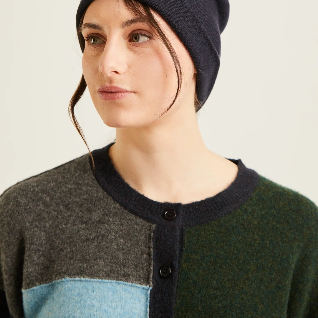 A woman wearing a dark navy knit beanie and a color-blocked knit sweater with gray, dark green, and light blue panels, photographed against a plain light background.