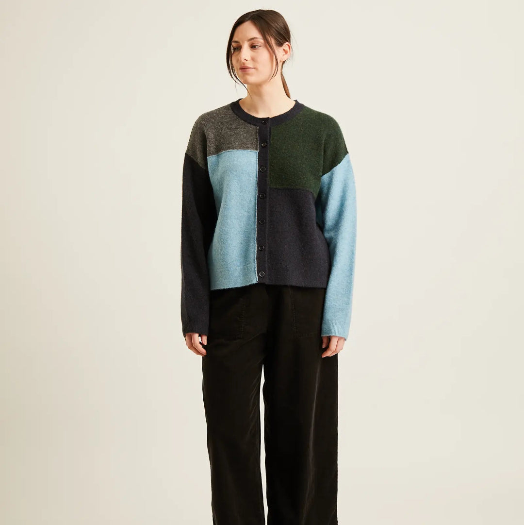 A woman stands against a plain light beige studio background wearing a color-block knit sweater with dark green, light blue, and charcoal panels, paired with wide-leg black pants and black flats.