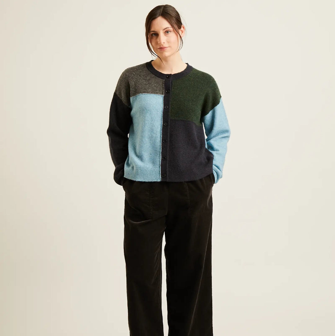 A woman stands against a plain light beige studio background wearing a color-block knit sweater with dark green, light blue, navy, and gray panels, black wide-leg corduroy pants, and black flats.