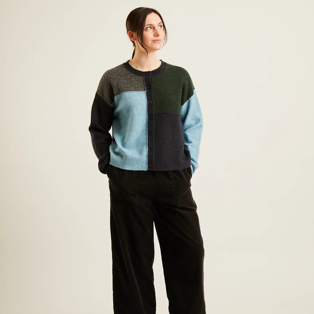 A woman modeling a color-block knit sweater with dark green, light blue, and black panels, paired with black wide-leg trousers and black flats, against a plain light beige studio background.