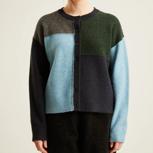 A woman wearing a color-block knit cardigan sweater with dark green, light blue, and dark gray panels, paired with black pants, against a plain light beige background.