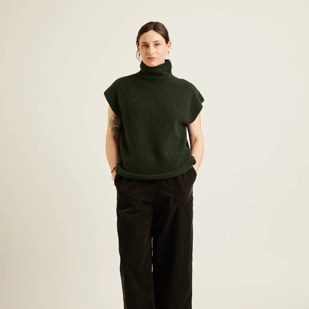 A model stands against a plain light beige studio background wearing a dark forest green short-sleeve turtleneck sweater and black wide-leg trousers.