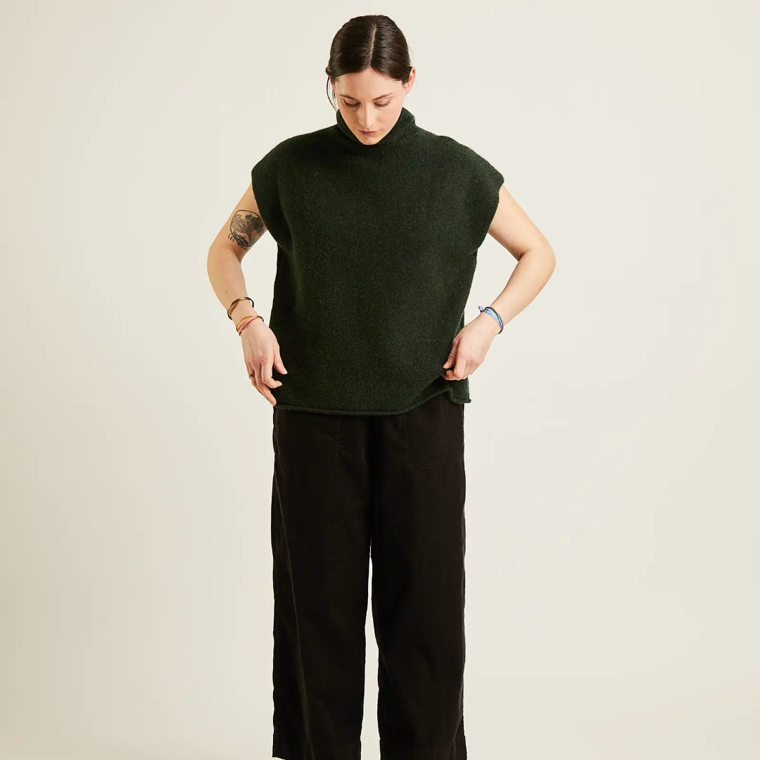 A model stands against a plain light beige studio background wearing a dark forest green sleeveless mock-neck knit sweater and black wide-leg trousers.