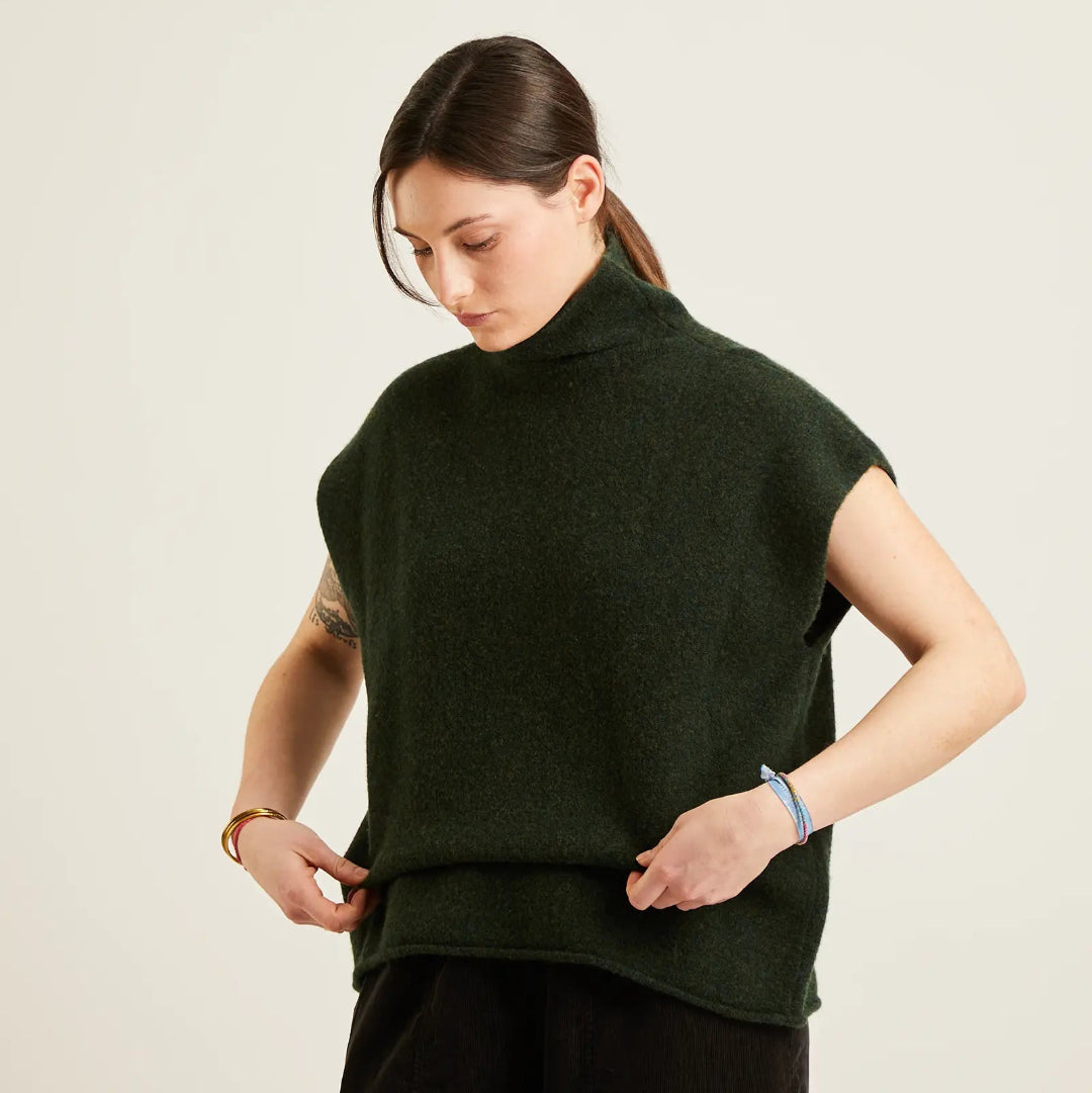 A woman wearing a dark forest green sleeveless mock-neck knit sweater and black trousers, standing against a plain light beige background.