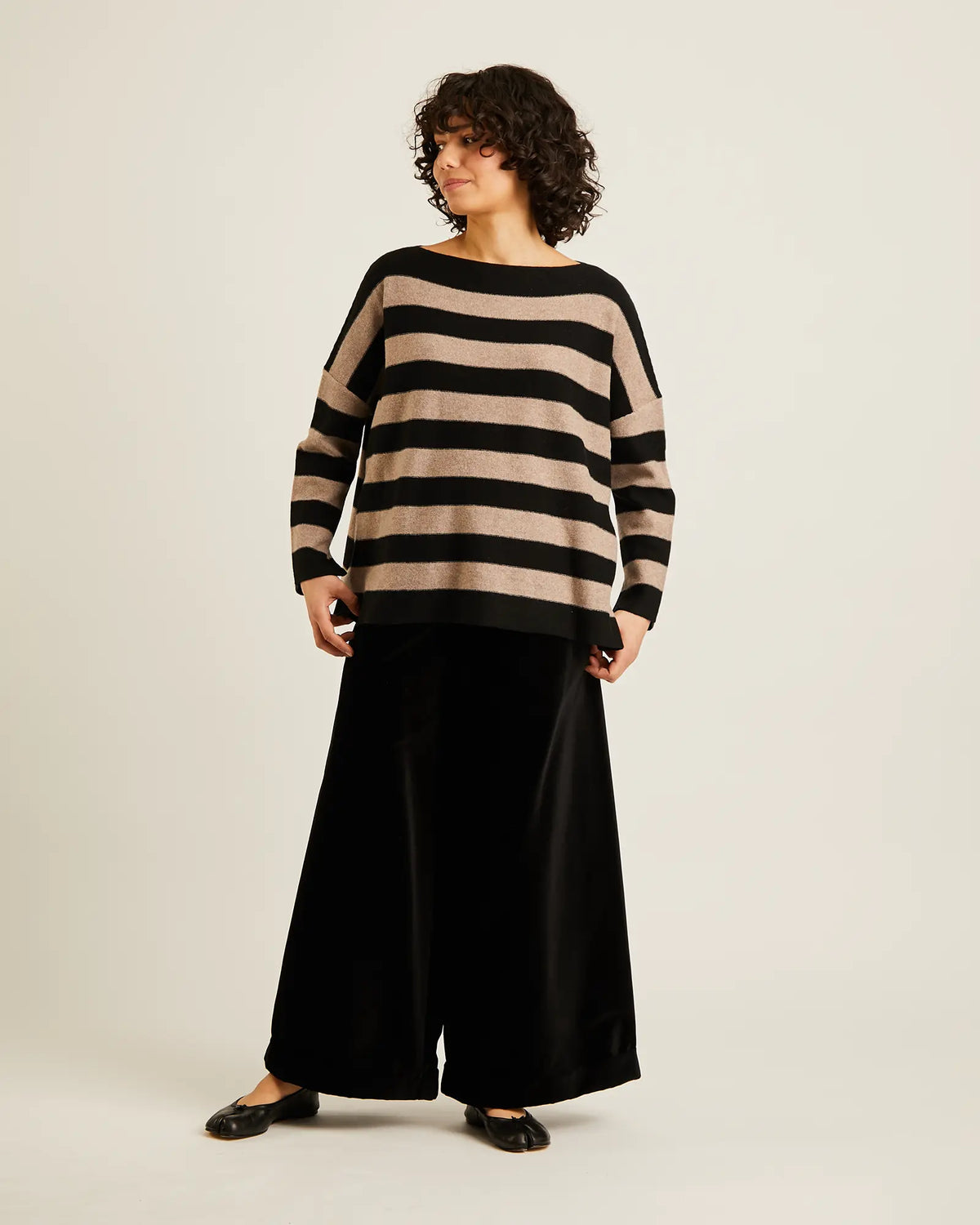 A woman modeling a black and beige horizontally striped knit sweater with wide boat neck and cuffs, paired with black wide-leg trousers and black flats, against a plain light beige studio background.