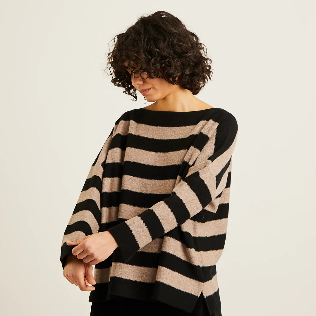 A woman wearing a loose, long-sleeve knit sweater with horizontal beige and black stripes, paired with black pants, photographed against a plain light background.