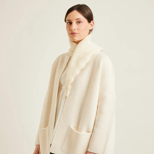 A woman modeling a cream-colored textured knit cardigan and a matching scalloped scarf, paired with black trousers, against a plain light beige studio background.