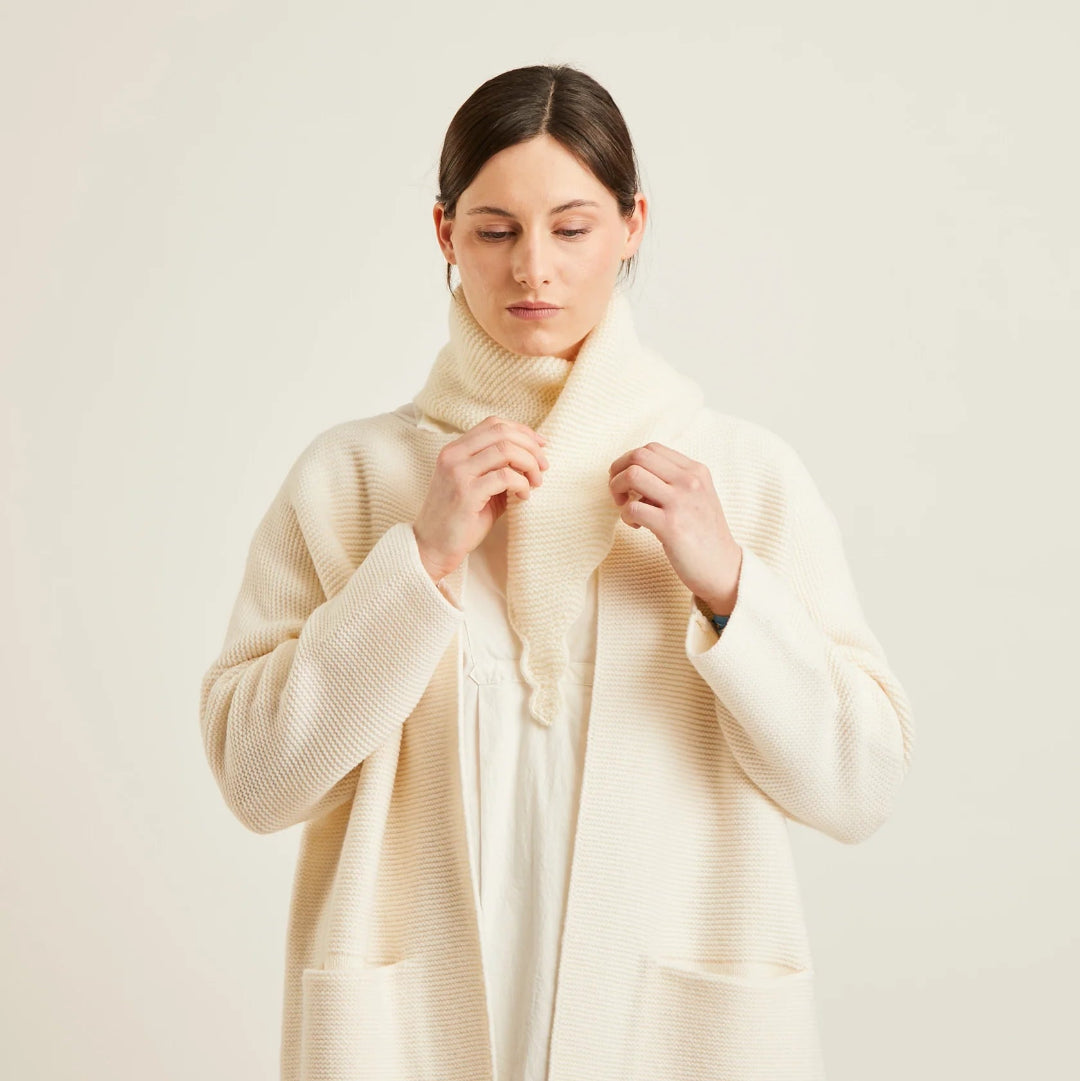 A woman wearing a cream-colored ribbed knit cardigan with a matching knit scarf, styled over a white top and black pants, against a plain light beige background.