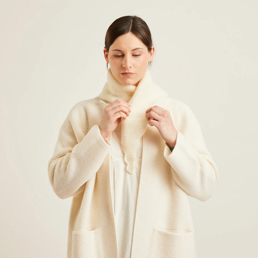 A woman wearing a cream-colored ribbed knit cardigan with a matching knit scarf, styled over a white top and black pants, against a plain light beige background.