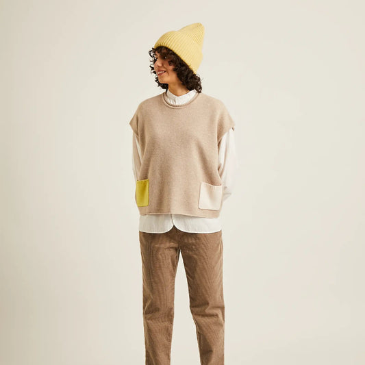 A model stands against a light beige studio background wearing a beige knit sweater vest with yellow and white color-blocked pocket details, a white mock-neck shirt, brown trousers, and a yellow beanie.
