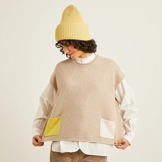 A model wearing a beige knit sweater vest with two color-blocked pockets (one yellow, one cream) over a white long-sleeve shirt, brown pants, and a yellow ribbed beanie, set against a plain off-white studio background.