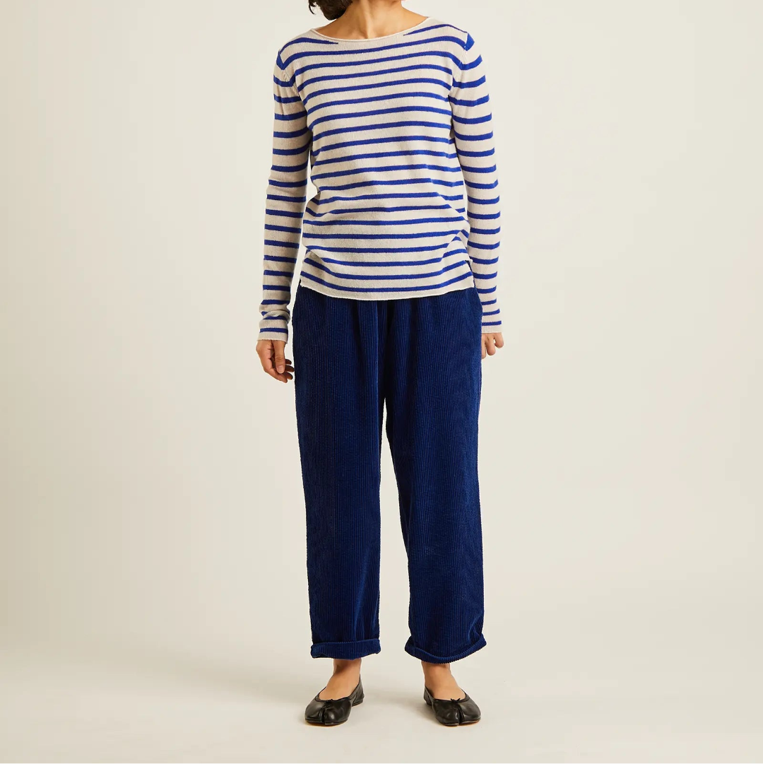 A model wearing a blue and white striped long-sleeve top and dark blue corduroy pants, standing against a plain light beige studio background.