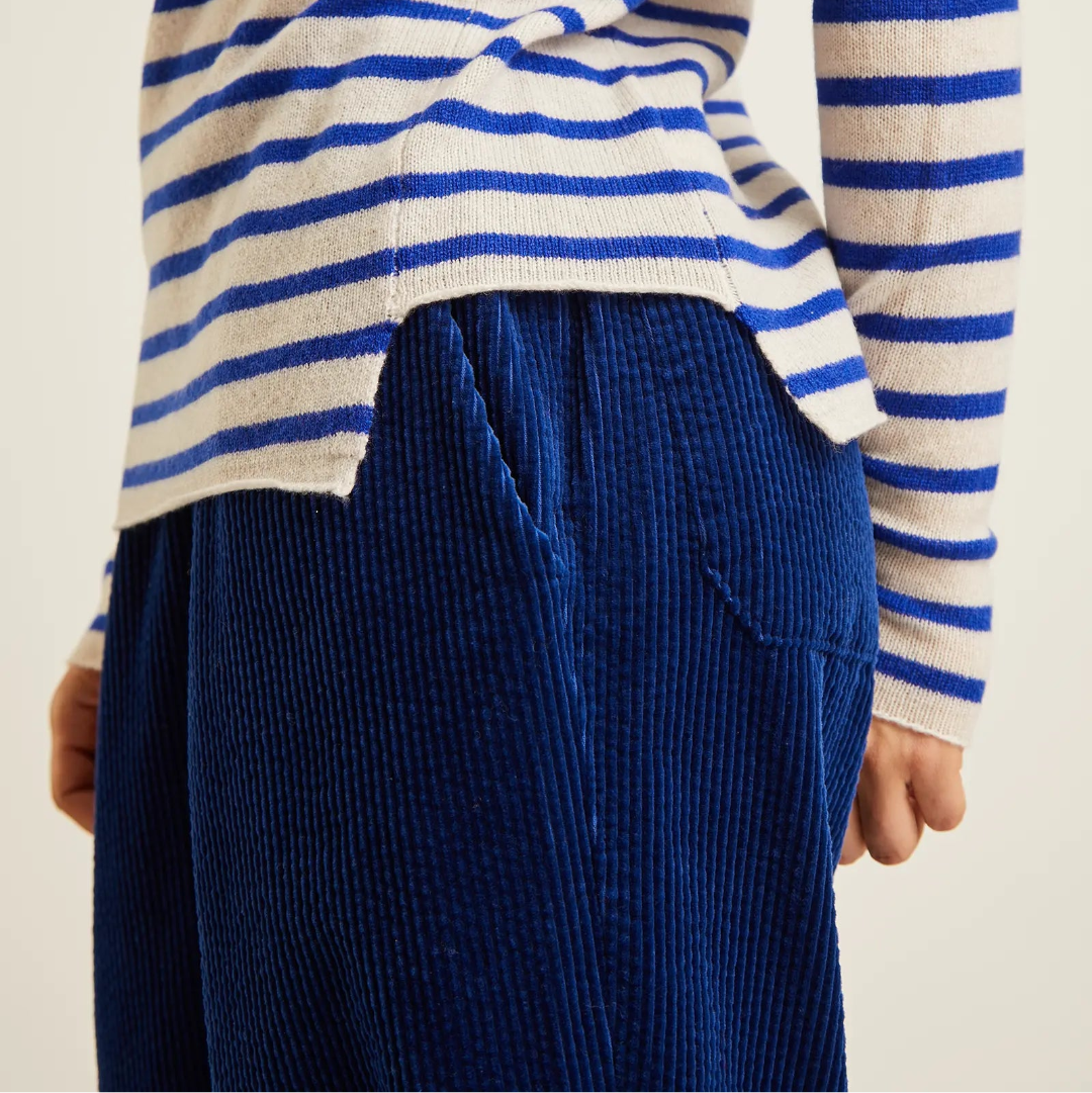 A close-up of a person wearing a blue and white striped knit top and dark blue corduroy trousers, shown from the chest down against a plain light background.