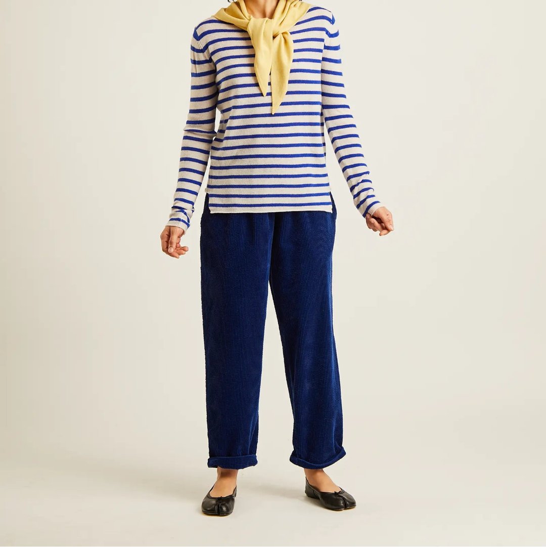 A woman modeling a blue and white striped long-sleeve top with a yellow neck scarf, paired with navy blue trousers and black flats, against a plain light beige studio background.