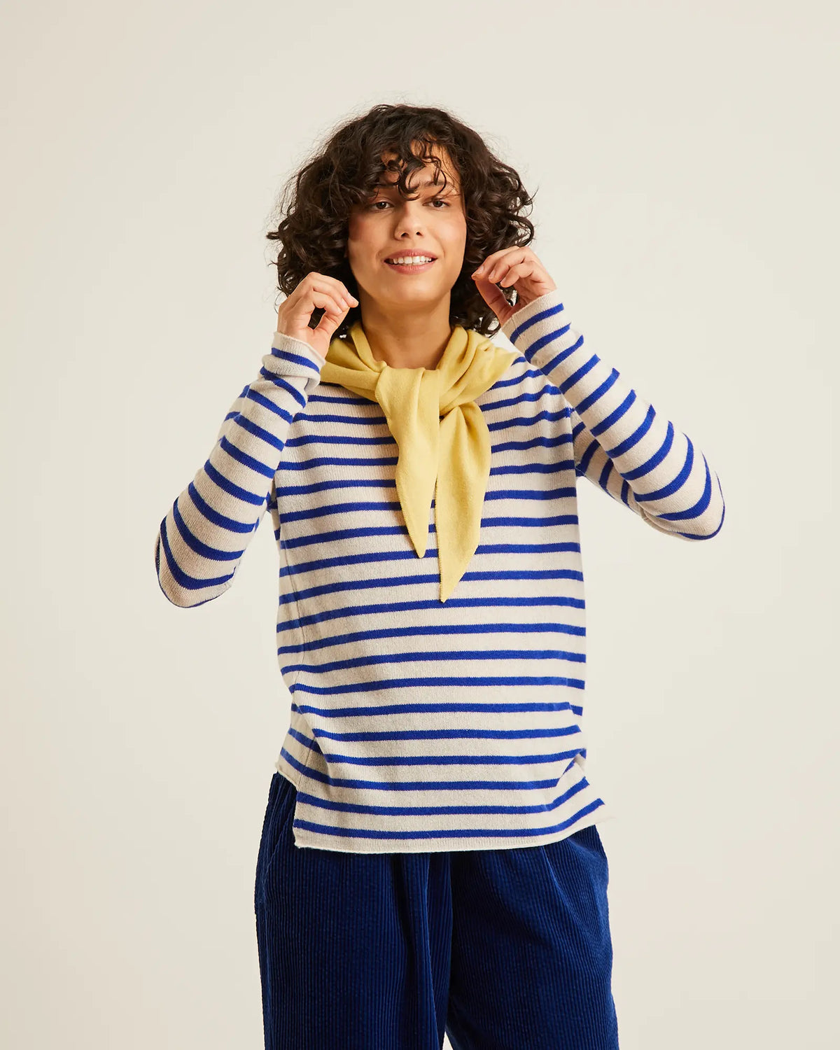 A woman with curly dark hair wearing a blue and white striped long-sleeve top, a yellow neck scarf, and dark blue corduroy pants, posing against a light beige studio background.