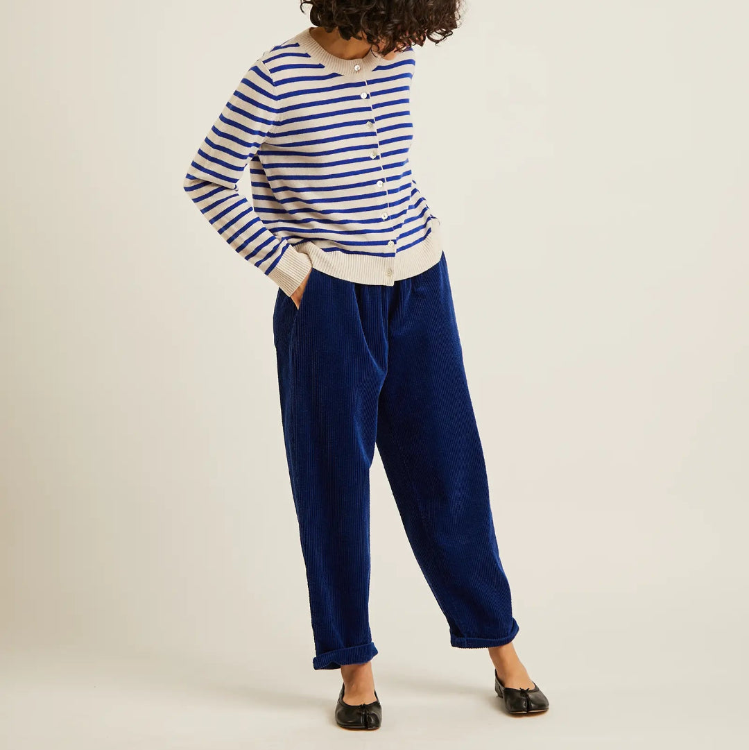 A model wearing a blue and white striped long-sleeve top, navy blue velvet trousers, and black flats, standing against a plain off-white studio background.