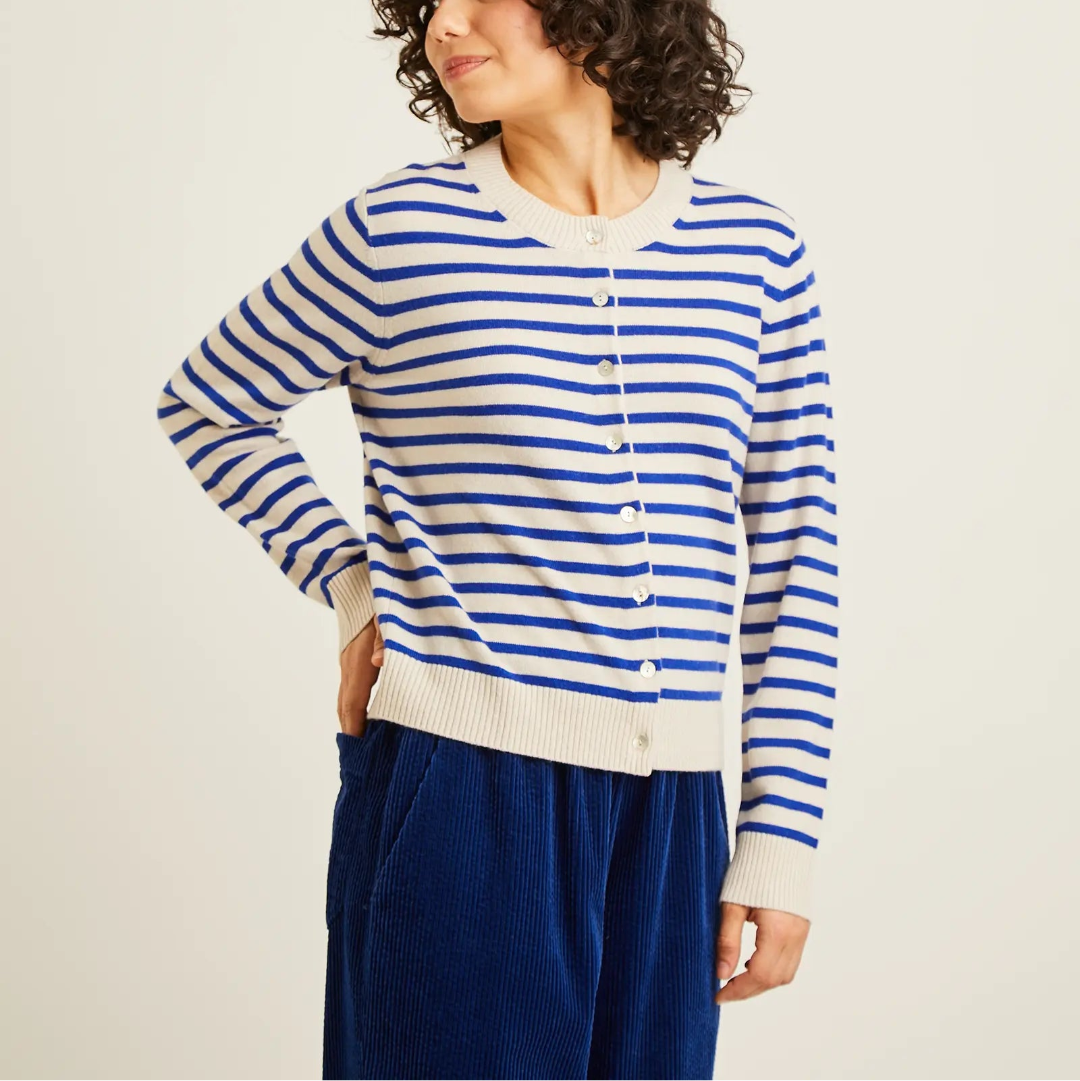 A woman with curly dark hair wearing a blue and cream striped button-up cardigan and dark blue corduroy trousers, posing against a plain off-white studio background.