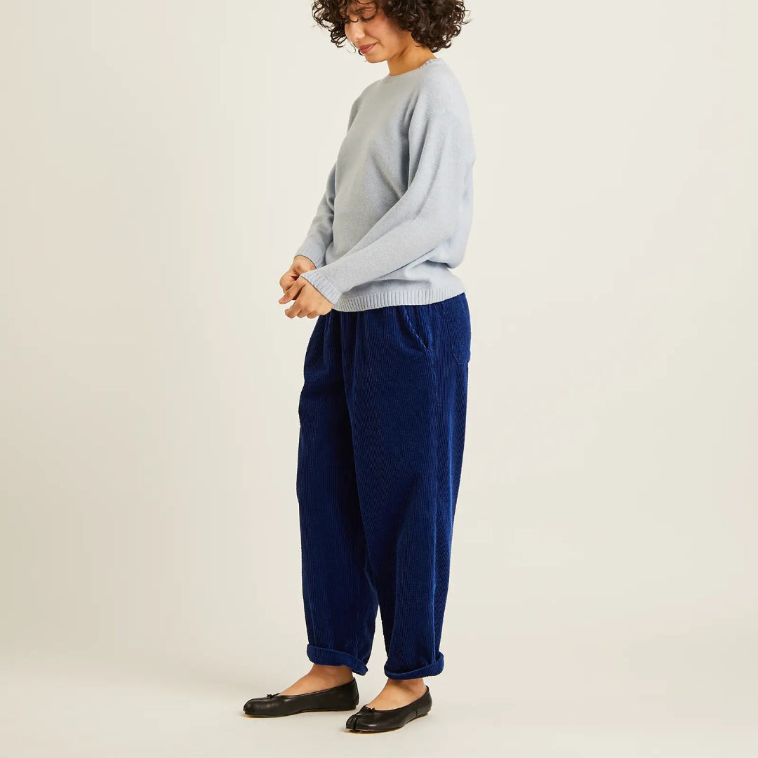 A model wearing a light blue knit sweater and navy blue corduroy pants, standing against a plain off-white studio background.