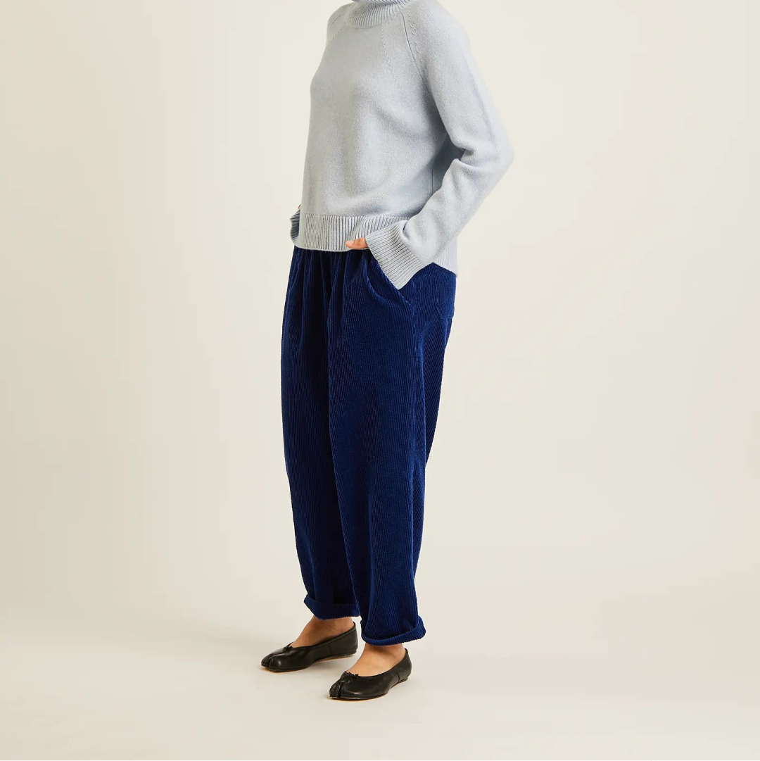 A woman stands in profile wearing a light blue turtleneck sweater, dark navy tapered corduroy pants, and black flats against a plain off-white studio background.