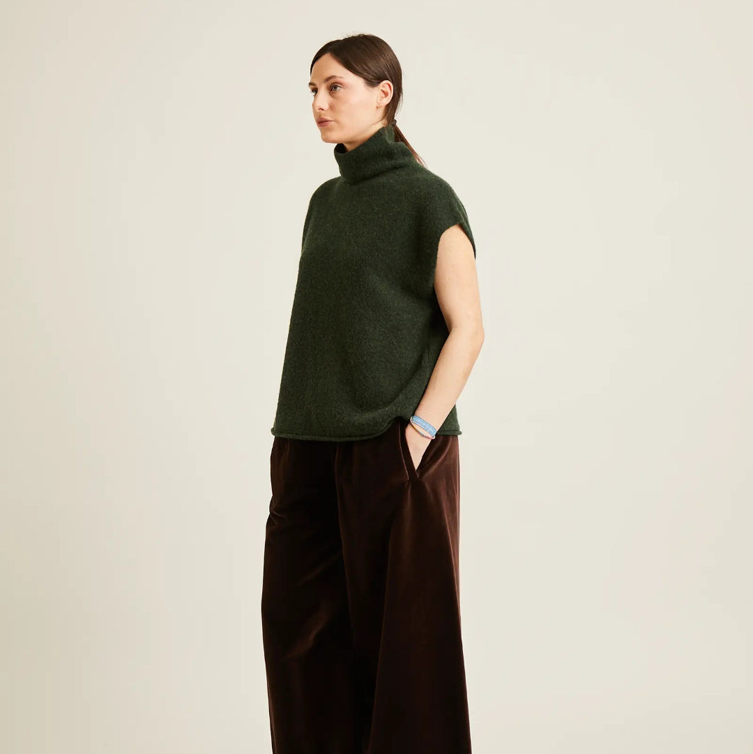 A woman stands in profile wearing a dark green sleeveless mock-neck knit top and dark brown wide-leg trousers against a plain light beige studio background.