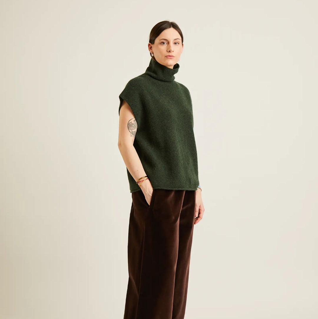 A model stands in profile wearing a dark green sleeveless mock-neck knit top and dark brown wide-leg trousers against a plain light beige studio background.